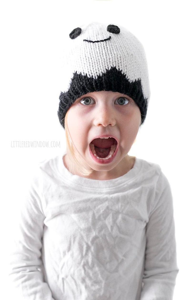 K&ouml;nnte beinhalten: Ein Kind tr&auml;gt eine wei&szlig;e und schwarze Strickm&uuml;tze mit einem l&auml;chelnden Gesicht. Die M&uuml;tze hat schwarze Augen und einen schwarzen Mund. Das Kind tr&auml;gt ein wei&szlig;es Hemd und hat den Mund in einem &uuml;berraschten Ausdruck ge&ouml;ffnet.