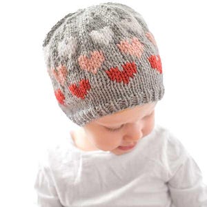 May include: A gray knitted hat with a pattern of ombre hearts in shades of pink and red. The hat is on a child's head.