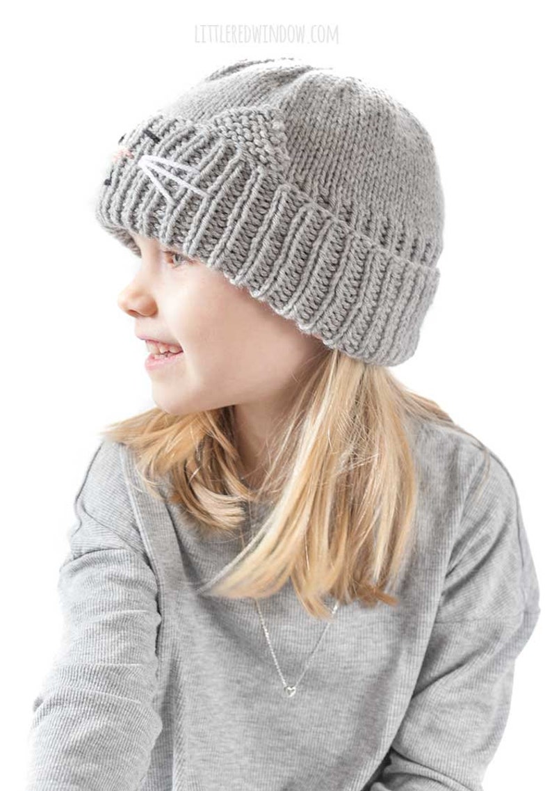 May include: A gray knitted beanie with a cat face embroidered on the front. The beanie is being worn by a child with long blonde hair.