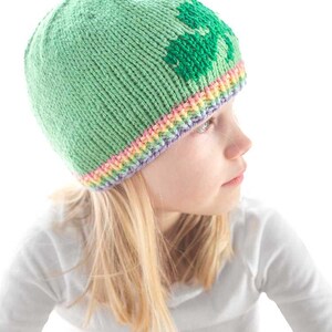 A child is sporting a knitted green shamrock hat, featuring a darker heart pattern and a multicolored rainbow brim. The child is facing sideways in a light gray shirt against the bright white background.