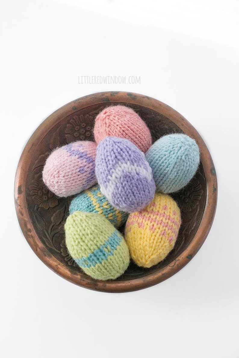 view from above of seven pastel colored knit Easter Eggs in various patterns in a dark brozne colored metal bowl on a white tabletop