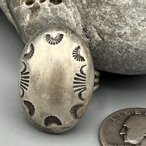 May include: An oval silver ring featuring a smooth, slightly tarnished surface with a series of stamped fan-shaped patterns around the perimeter. The ring is positioned next to a United States quarter coin for scale, showing its relative size.