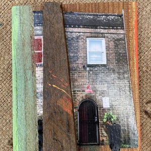 May include: A square wall art piece featuring a photograph of a brick building with a dark arched door, a single window, and a potted plant. The image is layered with vertical strips of weathered wood in shades of green, brown, and orange.