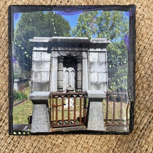 May include: A square slate coaster featuring a printed image of a stone mausoleum with Egyptian-style columns and a winged sun disk carving above the entrance. A rusted metal gate stands in front of the structure, surrounded by green trees.