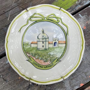 May include: A decorative white ceramic plate with a green scalloped edge and a central image of a building with a dome and the text "CHATEAU LATOUR". The plate has a green bow design and is held by metal clips.