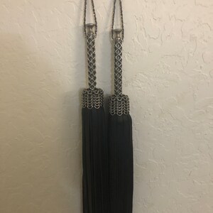 K&ouml;nnte beinhalten: Zwei schwarze Quasten-Vorhangbinder mit silbernen Kettenhemd-Oberteilen. Die Quasten h&auml;ngen an Haken an einer wei&szlig;en Wand.