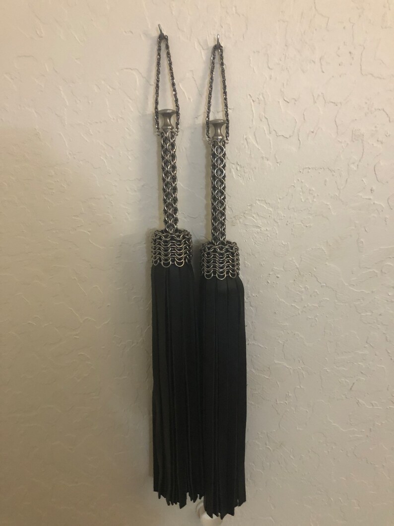 K&ouml;nnte beinhalten: Zwei schwarze Quasten-Vorhangbinder mit silbernen Kettenhemd-Oberteilen. Die Quasten h&auml;ngen an Haken an einer wei&szlig;en Wand.