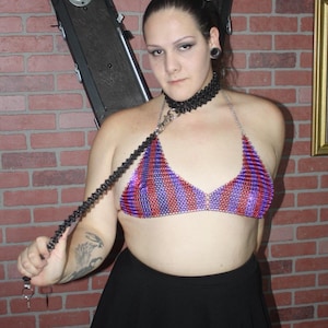 May include: A person wearing a black choker necklace and a purple and red striped chainmail bikini top. The person is holding a black chain leash.