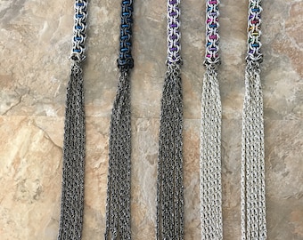 Könnte beinhalten: Sechs silberne Kettenpeitschen mit unterschiedlichfarbigen Griffen. Die Griffe bestehen aus Metallringen in einem Kettenhemdmuster. Die Peitschen haben mehrere Kettenstränge mit spitzen Enden.