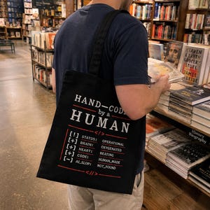 May include: Black tote bag with white text that reads "HAND-CODED by a HUMAN." The bag's design includes a code-like list with status updates. The person is in a bookstore, browsing books.