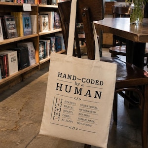 May include: A beige tote bag with the text "HAND-CODED by a HUMAN" in black. The bag's design includes a list of human attributes. The bag is hanging on a wooden chair in a bookstore.
