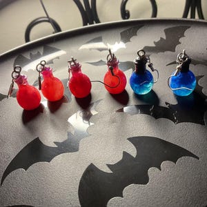 May include: A collection of potion bottle earrings in shades of red and blue. The earrings are displayed on a dark gray plate with bat silhouettes. Each bottle has a silver cap and hook.