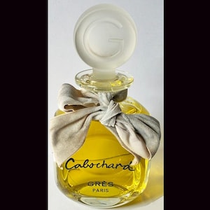 May include: A vintage perfume bottle filled with yellow liquid. The bottle has a clear, round stopper with a frosted "G" on top. A gray ribbon is tied around the neck. The label reads "Cabochard" and "GRES PARIS".