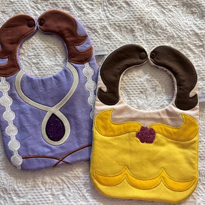 May include: Two baby bibs with princess themes. One bib is purple with a white necklace and a large purple gem. The other bib is yellow with a brown rose and a yellow dress. Both bibs have brown hair.