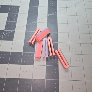 May include: A collection of pink and lavender U-shaped silicone tools, possibly for crafting or DIY projects. The tools are arranged on a gray and white checkered surface, with varying shades of pink and lavender.