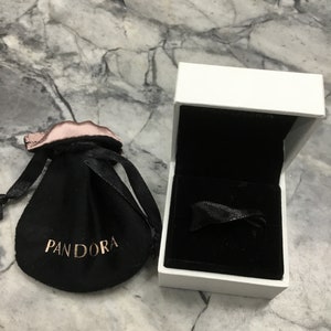 Puede incluir: Una bolsa de terciopelo negro con la palabra "Pandora" impresa en letras doradas, y una caja blanca con un interior de terciopelo negro.