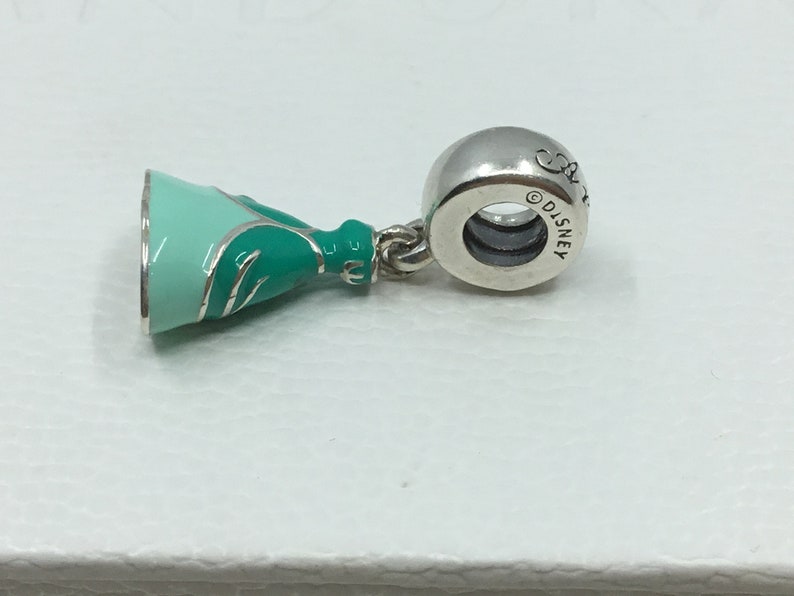 Puede incluir: Encanto de plata con un vestido de princesa verde y azul con ribete plateado. La parte posterior del encanto tiene la palabra "Disney" grabada.