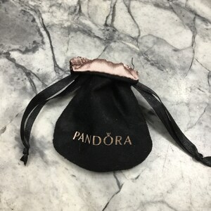 Puede incluir: Bolsa de terciopelo negro con cord&oacute;n de sat&eacute;n rosa y la palabra "PANDORA" impresa en letras doradas.