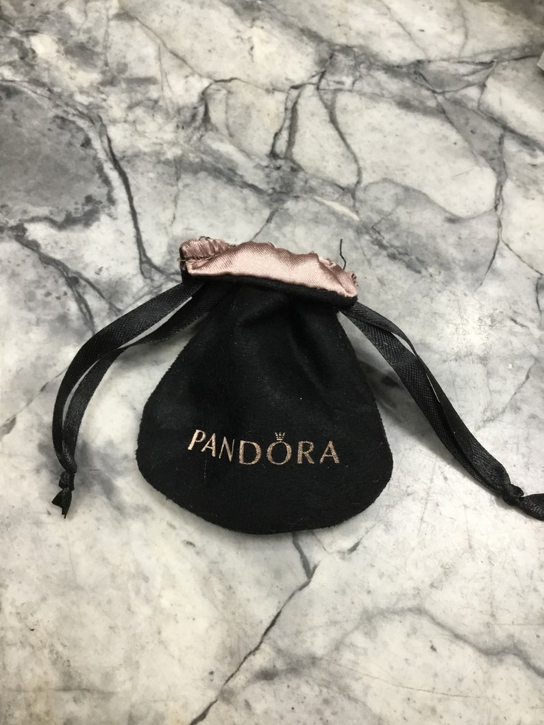 Puede incluir: Bolsa de terciopelo negro con cord&oacute;n de sat&eacute;n rosa y la palabra "PANDORA" impresa en letras doradas.