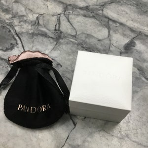 Puede incluir: Una bolsa de terciopelo negro con la palabra "PANDORA" impresa en letras doradas. La bolsa est&aacute; sobre una superficie de m&aacute;rmol blanco junto a una caja blanca con la palabra "PANDORA" impresa en la parte superior.