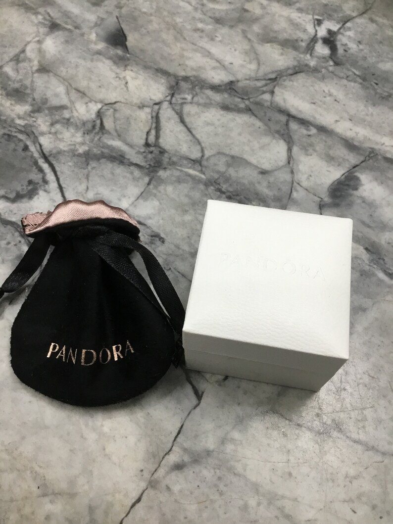 Puede incluir: Una bolsa de terciopelo negro con la palabra "PANDORA" impresa en letras doradas. La bolsa est&aacute; sobre una superficie de m&aacute;rmol blanco junto a una caja blanca con la palabra "PANDORA" impresa en la parte superior.