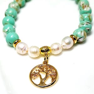 May include: A green and white beaded bracelet with a gold tree of life charm. The bracelet is made with round beads and has a pearl accent.