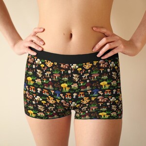 May include: Black boy shorts featuring a colourful all-over print of various mushrooms and green foliage. The underwear has a solid black elastic waistband and a fitted silhouette, showcasing a detailed pattern of yellow, brown, and red fungi.
