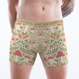 Könnte beinhalten: Boxershorts mit cremefarbenem Hintergrund und einem sich wiederholenden Muster aus roten und orangefarbenen Pilzen, grünen Farnen und Eichenblättern. Der beigefarbene Bund zeigt den Text "LIFE IS ANTIQUE, AND THE OLDEST ART". Der Stoff ist eng anliegend.