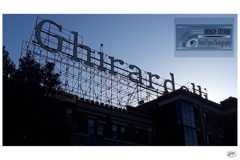 May include: A photograph of the Ghirardelli sign against a blue sky. The sign is large and white, with the word "Ghirardelli" prominently displayed. The sign is mounted on a metal framework above a brick building. The image is taken during the day.