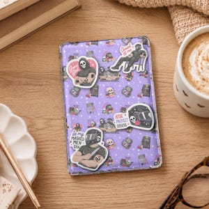 May include: A rectangular tablet case with a light purple background featuring a repeating pattern of skulls, books, and helmets. Three distinct stickers are applied to the surface, depicting masked figures, a motorcycle helmet, and text.