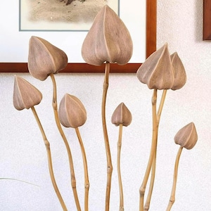 May include: A decorative wooden sculpture featuring multiple stems with light brown, petal-shaped tops. The stems are attached to a rounded, brown stone base. The sculpture is displayed on a white surface, with a potted plant and framed artwork in the background.