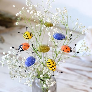 May include: A small, clear glass vase holds a bouquet of white baby's breath and colorful ladybug ornaments. The ladybugs are yellow, orange, and blue with black details. The vase sits on a light-colored wooden surface.