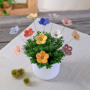 Könnte beinhalten: Ein weißer Keramiktopf mit einer dichten grünen Pflanze, die mit mehreren kleinen, bunten Keramikblumen an dünnen Metallstielen verziert ist. Die Blüten sind in den Farben Blau, Gelb, Orange, Rot, Rosa, Weiß und Violett gehalten.