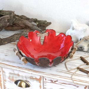 Peut inclure: Un bol en céramique rouge fait à la main, de forme organique unique. L'intérieur du bol est rouge brillant et l'extérieur est brun foncé texturé. Il est orné de poignées en bois et de détails en corde, créant une esthétique rustique.