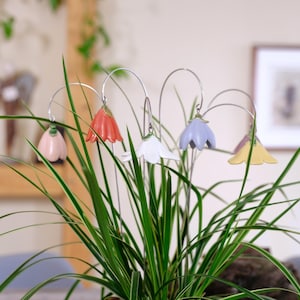 Könnte beinhalten: Eine Sammlung von sechs blumenförmigen Keramikornamenten in den Pastellfarben Rosa, Orange, Weiß, Hellblau, Violett und Gelb, die jeweils an einem dünnen, gebogenen silberfarbenen Metalldraht hängen und in einer grünen Topfpflanze stecken.