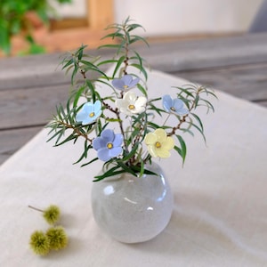 Peut inclure: Un petit vase en céramique gris clair de forme ronde contenant un bouquet de branches feuillues vertes agrémenté de fleurs en céramique à cinq pétales dans des tons bleu pâle, blanc et jaune doux, avec un petit centre texturé foncé.