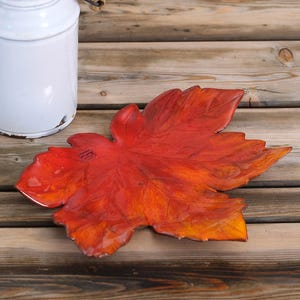 Könnte beinhalten: Eine keramikförmige Schale in Form eines Blattes in Rot- und Orangetönen, die an Herbstlaub erinnert. Die Schale liegt auf einer Holzoberfläche, im Hintergrund ist ein weißer Keramikbehälter zu sehen. Die Schale ist etwa 30 cm lang.