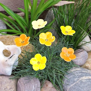 Könnte beinhalten: Eine Ansammlung von sechs Keramikblumen in leuchtenden Gelb- und Orangetönen, jede mit fünf abgerundeten Blütenblättern und einer dunklen Mitte. Die Blumen sind auf dünnen Metallstielen befestigt und zwischen grünem Gras, glatten Steinen und einem weißen Felsen angeordnet.