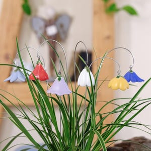 Peut inclure: Un ensemble de six tuteurs de jardin décoratifs avec des fleurs en céramique en forme de clochettes, déclinées en bleu clair, rouge, lavande, blanc, jaune et bleu foncé. Chaque fleur est suspendue à une tige métallique argentée et courbée.