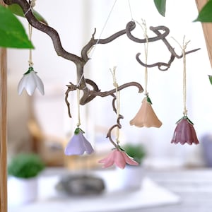 Könnte beinhalten: Fünf blumenförmige Keramikornamente hängen an einem gedrehten Ast. Jedes Ornament hat eine einzigartige Pastellfarbe, darunter Weiß, Lavendel, Rosa, Pfirsich und Mauve, mit einem kleinen grünen Blattdetail oben und einer dünnen beigen Schnur.