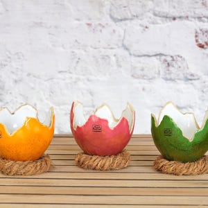 Może przedstawiać: Trzy dekoracyjne ceramiczne miski w kolorze pomarańczowym, czerwonym i zielonym, każda z unikalnym wzorem pękniętej skorupki jajka. Miski mają białe wnętrze i stoją na podkładkach z plecionej liny.