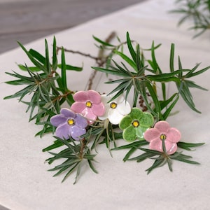 Könnte beinhalten: Fünf kleine Keramikblumen in den Farben Rosa, Weiß, Violett und Grün, jede mit einer gelben Mitte. Die Blumen sind zwischen dünnen, nadelartigen grünen Blättern auf einer hellen Stoffoberfläche angeordnet und bilden ein zartes Arrangement.