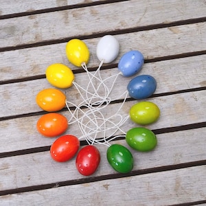 Puede incluir: Una colección de huevos de Pascua de madera pintados de colores, cada uno con una cuerda blanca adjunta. Los huevos están dispuestos en un patrón circular sobre una superficie de madera envejecida, mostrando una variedad de colores brillantes.