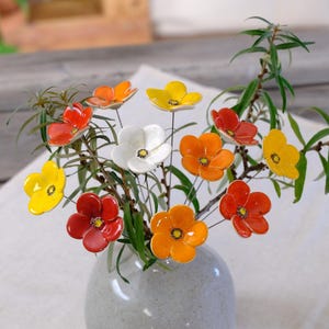 Könnte beinhalten: Eine Sammlung von Keramikblumen in leuchtenden Rot-, Orange-, Gelb- und Weißtönen, jede mit einer dunklen Mitte. Diese glänzenden, bunten Blüten sind an dünnen Metallstielen befestigt und in einer hellgrauen Keramikvase arrangiert.