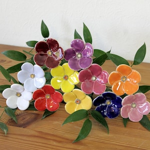 May include: A collection of eleven ceramic flowers in various colours including red, purple, white, yellow, orange, pink, and blue, each featuring five rounded petals and a small textured centre, arranged among several green leaves.