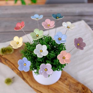 Pode incluir: Um conjunto de doze flores de cerâmica em várias cores pastel, cada uma com um centro amarelo e uma haste de metal fina. As flores estão dispostas num vaso branco cheio de folhagem verde, criando uma exibição vibrante de flores.