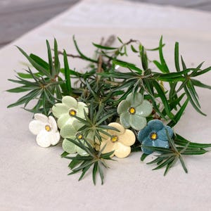 Op de afbeelding: Een verzameling kleine, vijfbladige keramische bloemen in tinten wit, lichtgroen, geel en blauw, elk met een geel hartje. Deze decoratieve keramische bloesems worden afgewisseld met dunne, puntige groene bladeren aan slanke stengels.