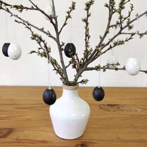 Könnte beinhalten: Eine weiße Keramikvase hält Zweige mit kleinen grünen Knospen und hängenden dekorativen Eiern. Die Eier sind schwarz und weiß und werden an dünnen Schnüren aufgehängt. Die Vase steht auf einer Holzoberfläche.