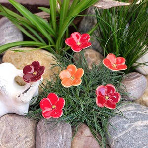 Könnte beinhalten: Ein Set aus sechs Keramikblumen-Ornamenten in Rot-, Orange- und tiefen Burgundertönen. Jede Blume hat fünf abgerundete Blütenblätter mit glänzender Oberfläche und einer kleinen gelben Mitte, befestigt an dünnen Drahtstielen zur Dekoration.