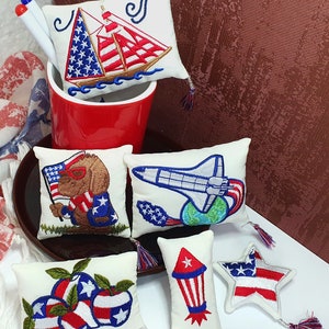 May include: A collection of small, white decorative pillows with patriotic embroidery. Designs include a sailboat, a dog with sunglasses holding a flag, a space shuttle, apples, a rocket, and a star, all in red, white, and blue. The text "Other pillows available in our shop" is visible.